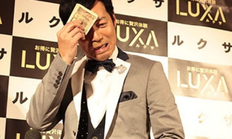 一万円札で汗を拭く定番ギャグを披露する東貴博、スーツ姿でコミカルな表情が印象的なワンシーン