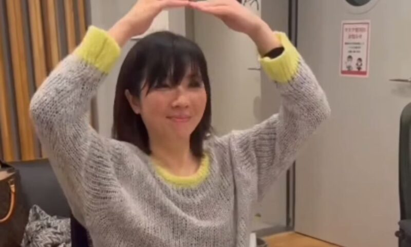 室内で両手を頭の上に掲げてポーズをとる岡本真夜。グレーのニットを着用し、リラックスした雰囲気で微笑むオフショット。