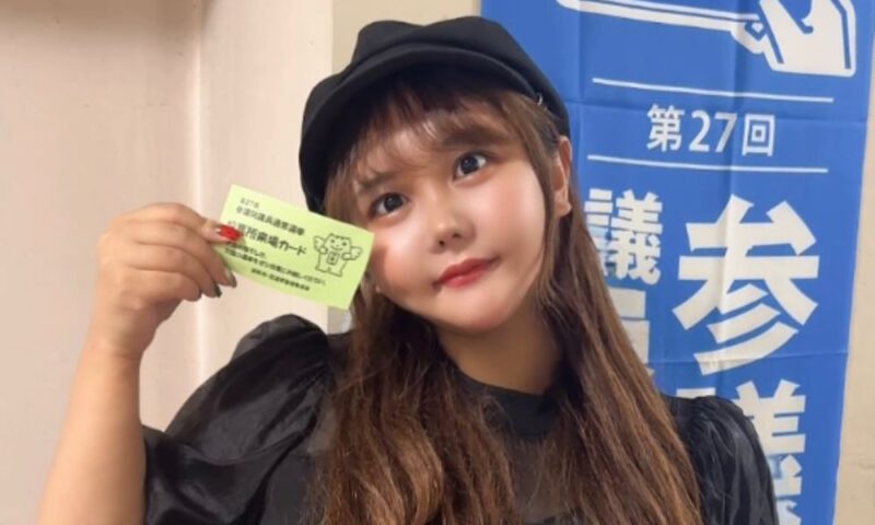 黒いベレー帽と黒のシースルー袖トップスを着たなな茶が、「期日前投票証明書」と書かれた緑色のカードを手に持ち、青い選挙ポスターの前でポーズをとっている様子