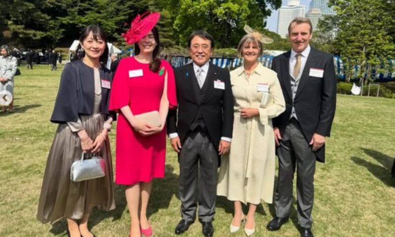 園遊会で赤いドレスと同色の帽子を着用した小野田紀美氏と、来場者たちが屋外で並んで撮影している様子