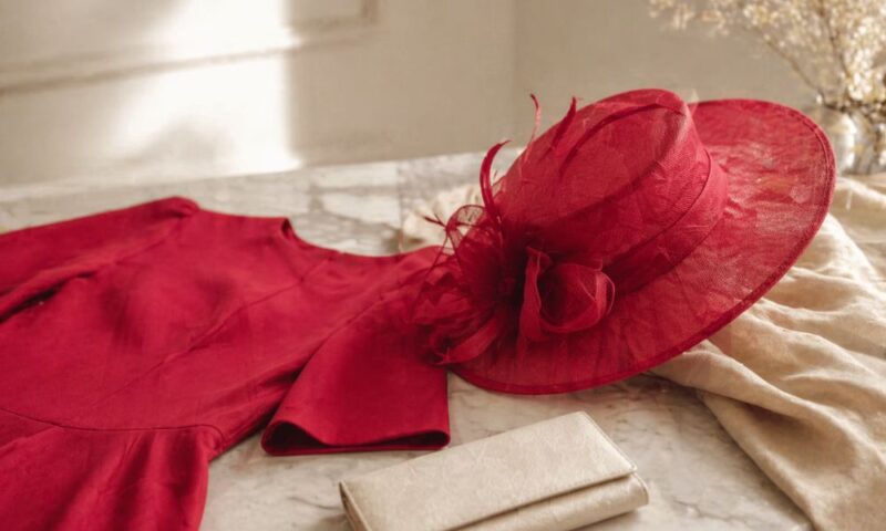 赤いドレスと同色の帽子、ベージュのクラッチバッグが大理石のテーブルの上に並べられたフォーマルコーディネートのイメージ