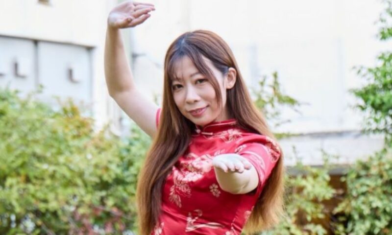 いぜん(芸人)が赤いチャイナドレスを着て、屋外でポーズを取りながら微笑んでいる様子