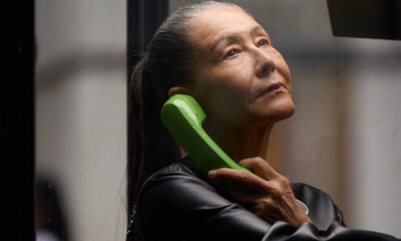 公衆電話の緑色の受話器を耳に当てる浅野順子。グレイヘアをポニーテールにした、横顔の近影写真