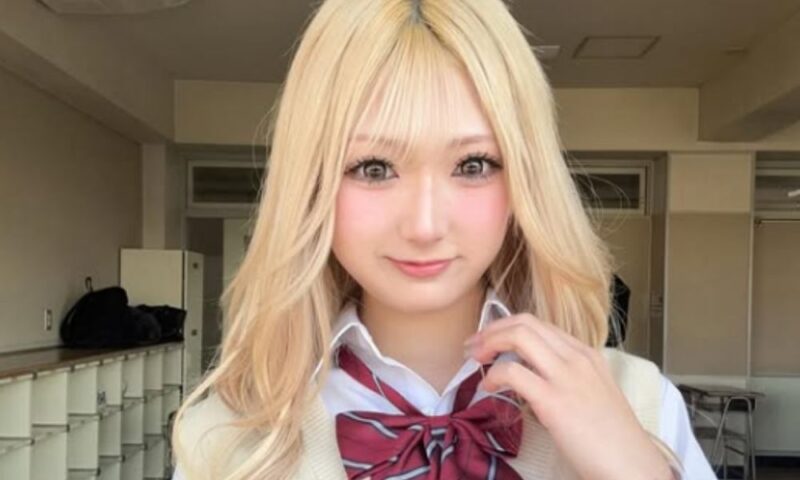 金髪のロングヘアで、白いシャツに赤いストライプのリボンを合わせた制服姿のじゅなさん。教室のような場所でカメラを見つめているショット