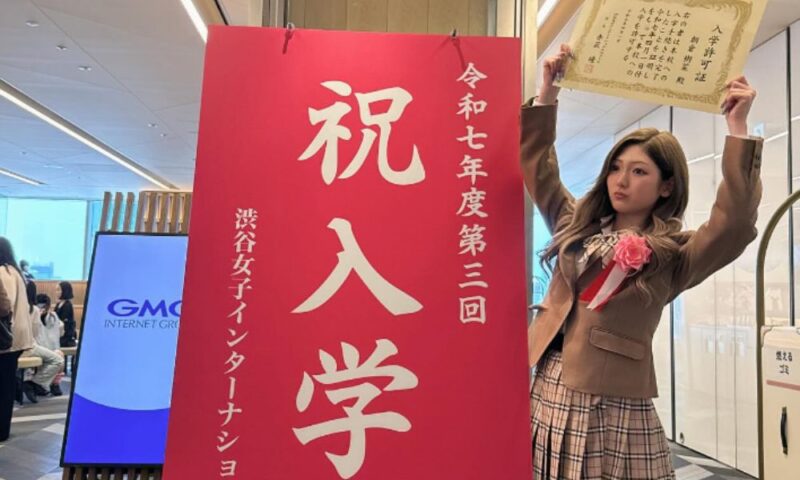 渋谷女子インターナショナルスクールの第三回入学式。赤い大きな看板には『令和七年度第三回 祝 入学』と書かれており、その横で、ベージュのジャケットとチェックのスカート姿のじゅな（朝倉樹菜）さんが、入学許可証を掲げて立つ様子。