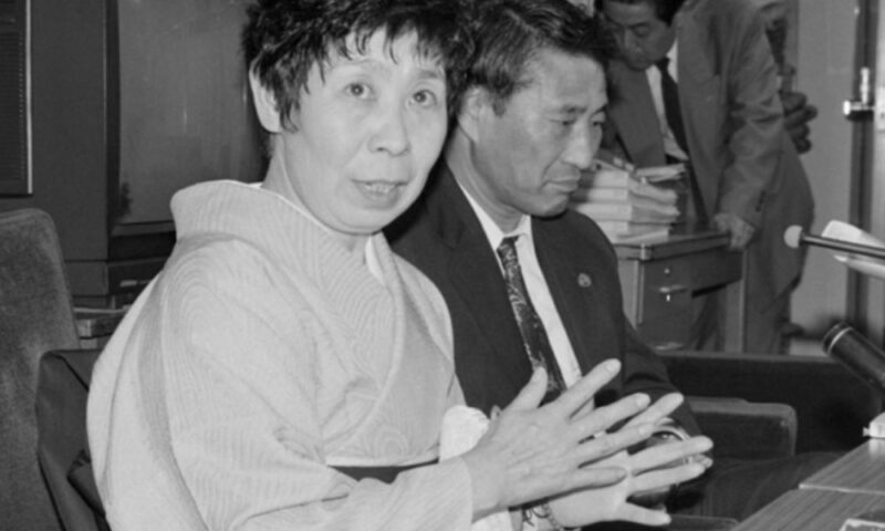 堀江ひとみさんが着物姿で話す昔の報道写真。机の前に座り、身振り手振りで説明している。隣にはスーツ姿の男性が座っている