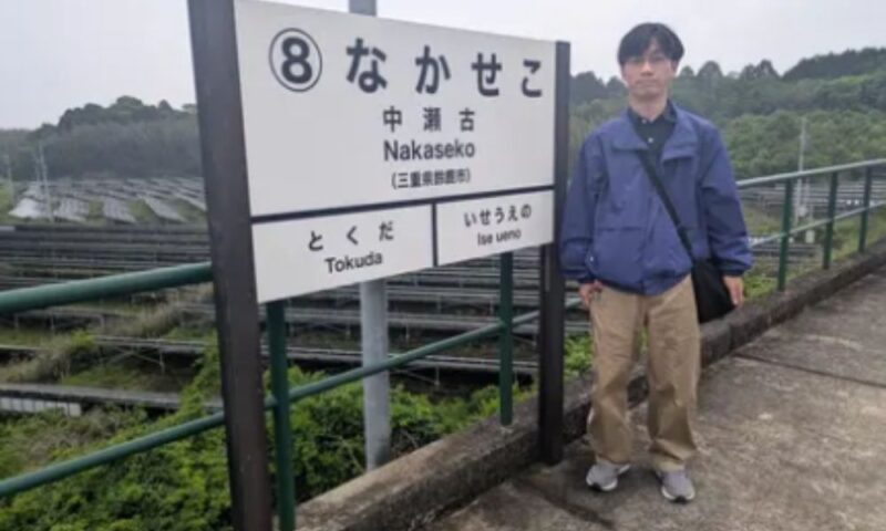 「なかせこ」と書かれた三重県の中瀬古駅の駅名標の横で、青いジャケットにベージュのパンツ姿の男性が立っている写真。背景には緑の斜面と線路が広がっている。