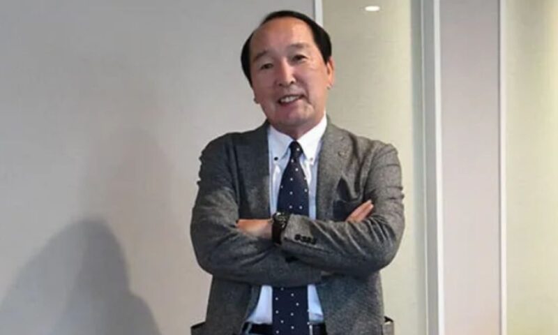 グレーのジャケットとネクタイを着た佐田繁理が、白い室内の壁の前で腕を組んで微笑んでいる写真