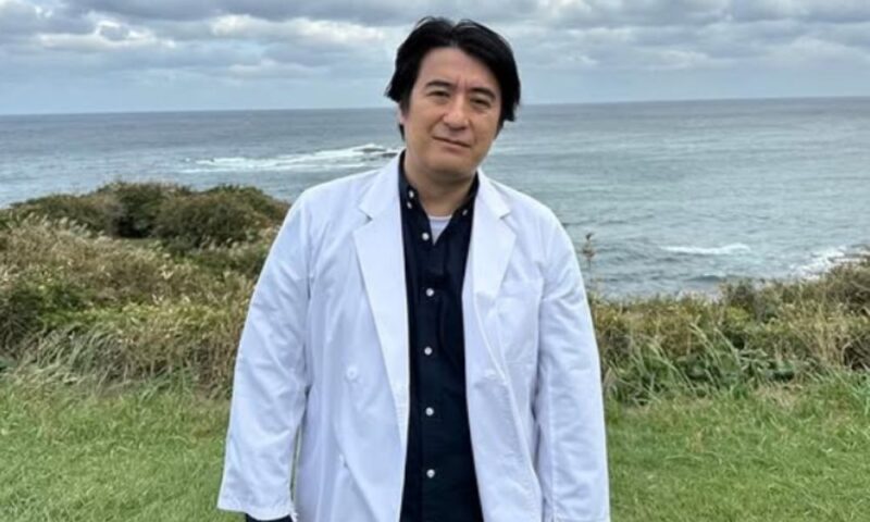 勝浦の海岸沿いの草地で白衣を着た男性が立っている写真。曇り空と太平洋の荒波が背景に広がる『佐久間宣行のオールナイトニッポン0(ZERO)』関連イメージ