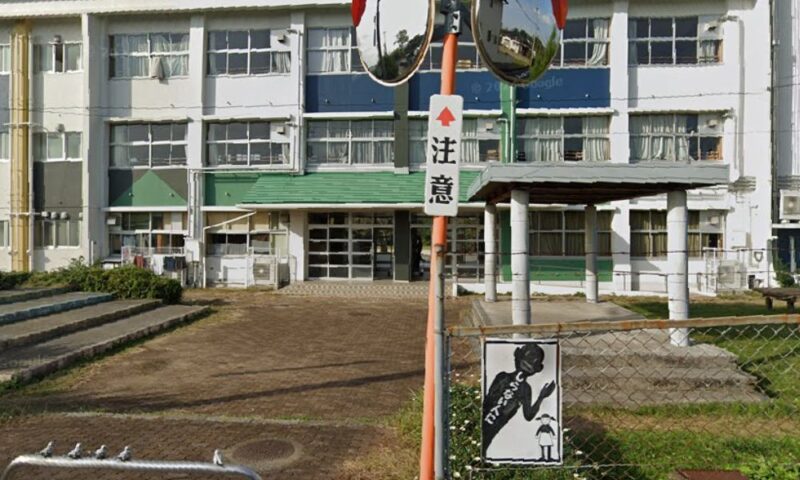 田川市立金川小学校の正面玄関付近を写した写真。3階建ての校舎と緑色の屋根の玄関が中央にあり、手前には「注意」と書かれた標識と、子どもの安全を呼びかけるイラスト看板、植え込みや通路が見えている。