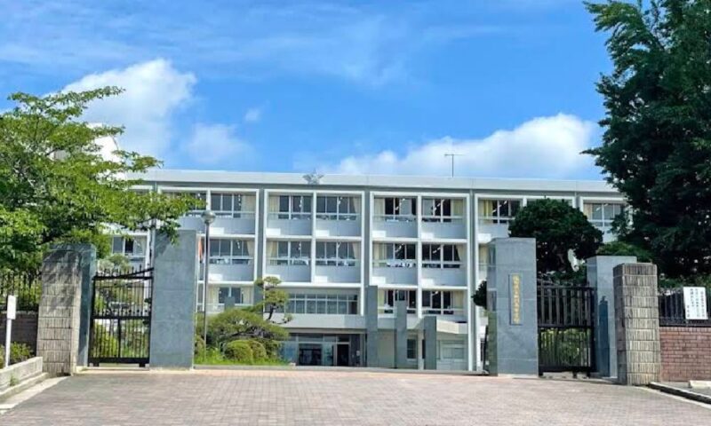 福岡県立田川高等学校の正門から校舎を望む写真。左右に石造りの門柱と黒い門扉があり、その奥に白い四階建ての校舎が建ち、青空と木々の緑に囲まれている。