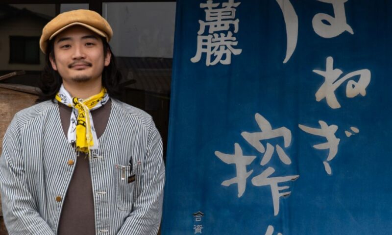 はねぎ搾りの酒蔵吉田屋5代目・吉田嘉一郎さん。藍色ののれんに「はねぎ搾り」「萬勝」と書かれた蔵の前で微笑む様子(長崎県南島原市)