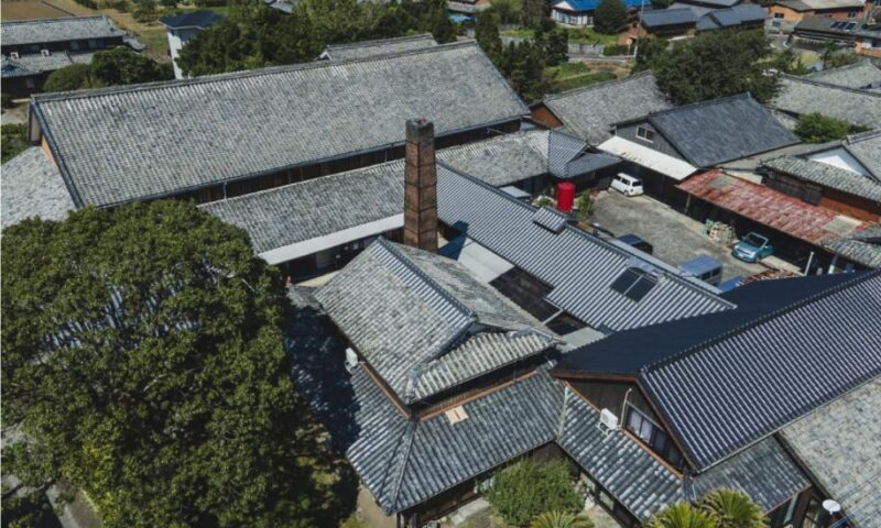 長崎県南島原市にあるはねぎ搾りの酒蔵・吉田屋の全景。灰色の瓦屋根が連なる伝統的な蔵と煉瓦造りの煙突が特徴的な敷地(空撮)